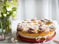 Torta di rose soffice e dorata, cosparsa di zucchero a velo, decorata con un nastro a cuoricini. Accanto, un vaso con fiori bianchi.