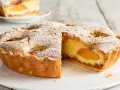 Una fetta di torta alle pesche, con la crosta dorata e un ripieno succoso, cosparsa di zucchero a velo e mandorle.