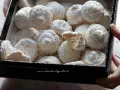 Scatola piena di soffici dolcetti al cocco, spolverati di zucchero a velo. Uno è spezzato, rivelando un interno friabile.