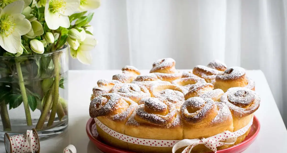 Torta di rose soffice e dorata, cosparsa di zucchero a velo, decorata con un nastro a cuoricini. Accanto, un vaso con fiori bianchi.