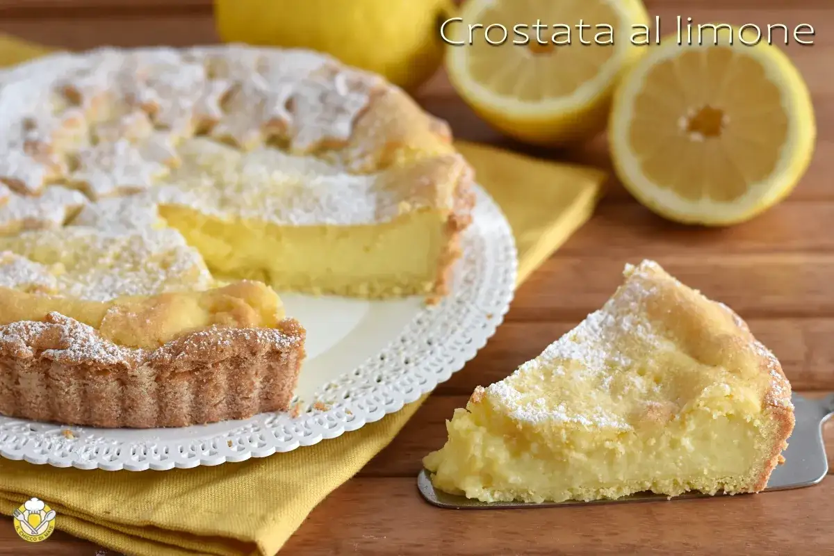 Una fetta di crostata al limone, cosparsa di zucchero a velo, su un piatto bianco. Sullo sfondo, la torta intera e spicchi di limone.